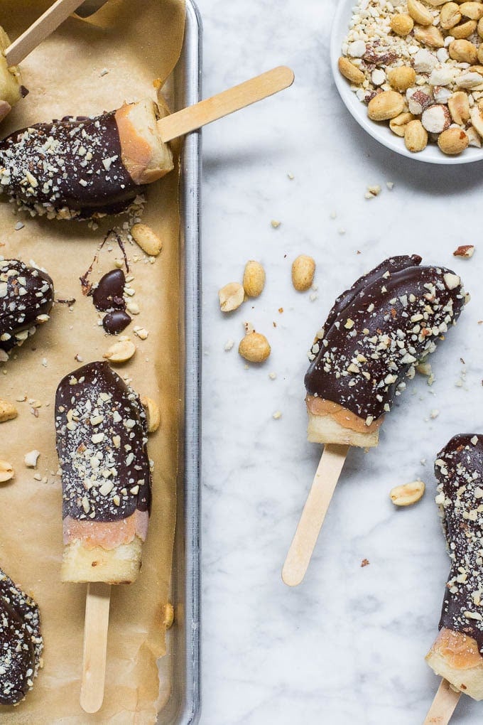 Aerial view of a baking sheet with brown parchment paper and frozen bananas covered in chocolate, peanut butter and roasted peanuts with a bowl of peanuts in the right upper corner.