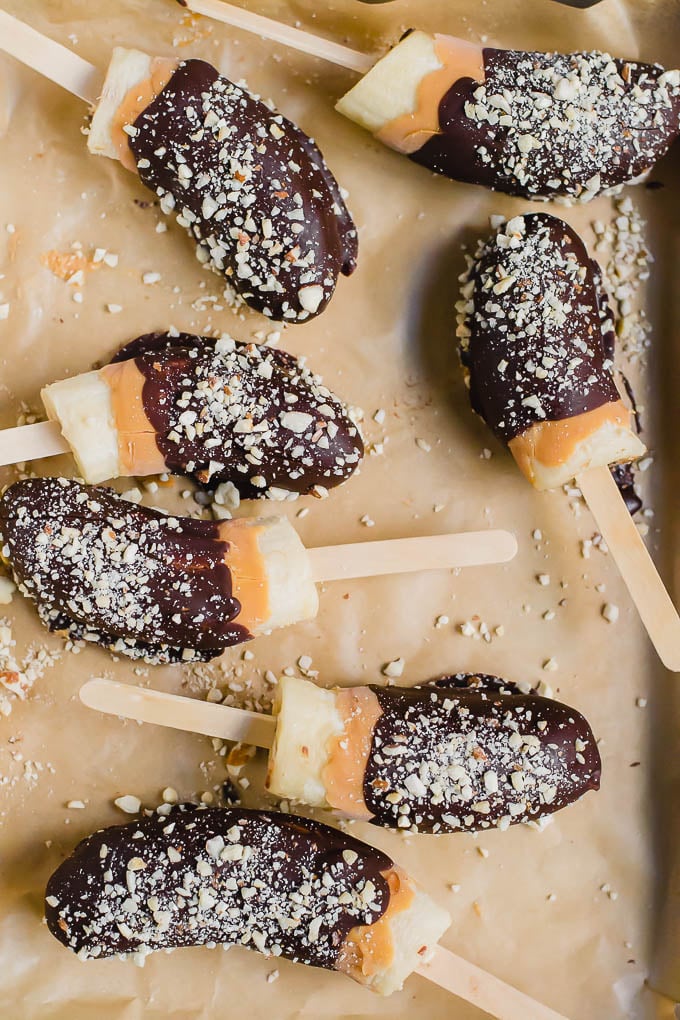 An aerial view of frozen banana pops covered with chocolate, peanut butter, and roasted peanuts on a brown piece of parchment paper. 
