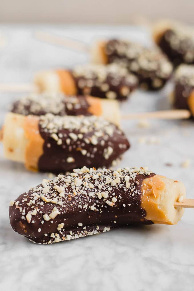 Straight on view of bananas on a stick covered in chocolate, peanut butter and roasted peanuts. 