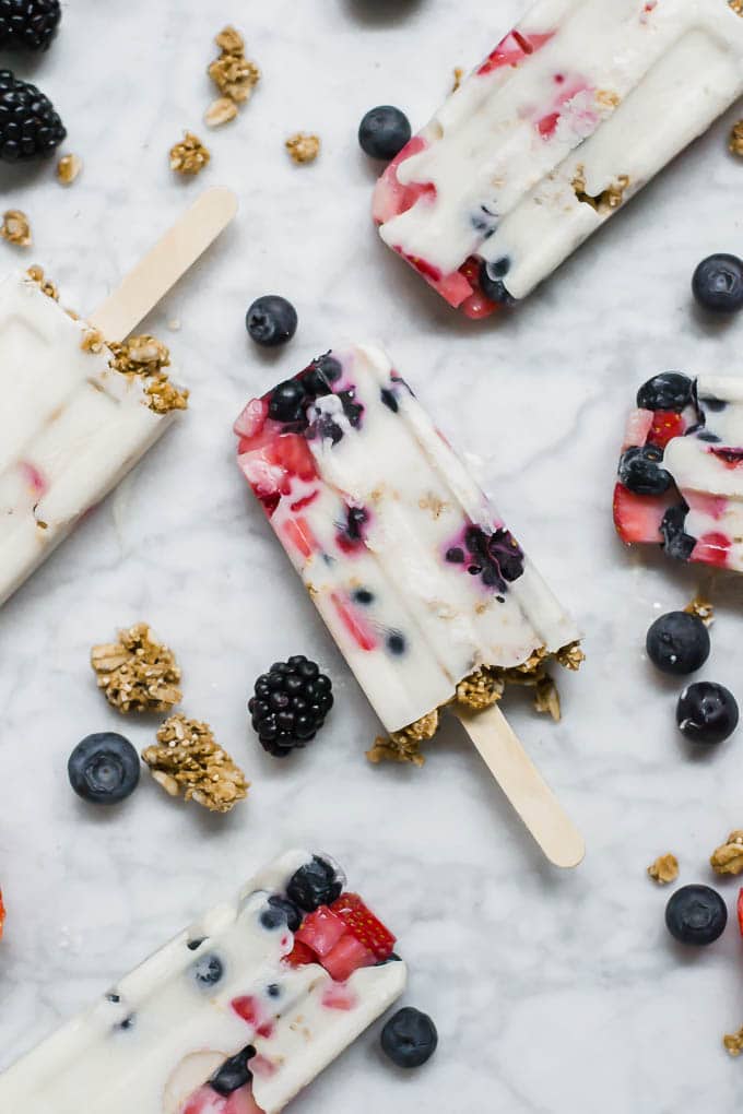 An aerial view of yogurt and granola parfait breakfast popsicles made with dairy-free yogurt and scattered with gluten-free granola and fresh berries on a marble surface. 