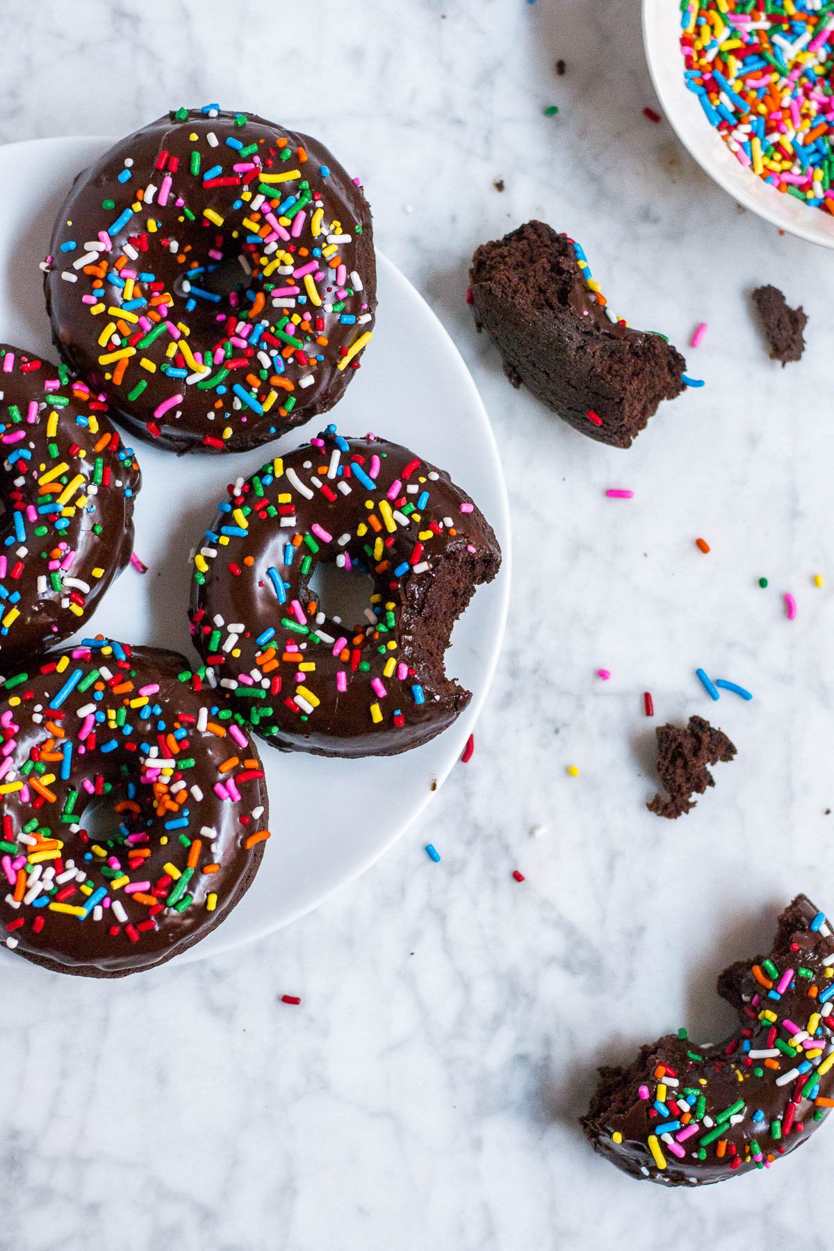 Aerial view of a plate with donuts and pieces of donuts on a marble table and a small bowl with sprinkles in the upper right hand corner. 