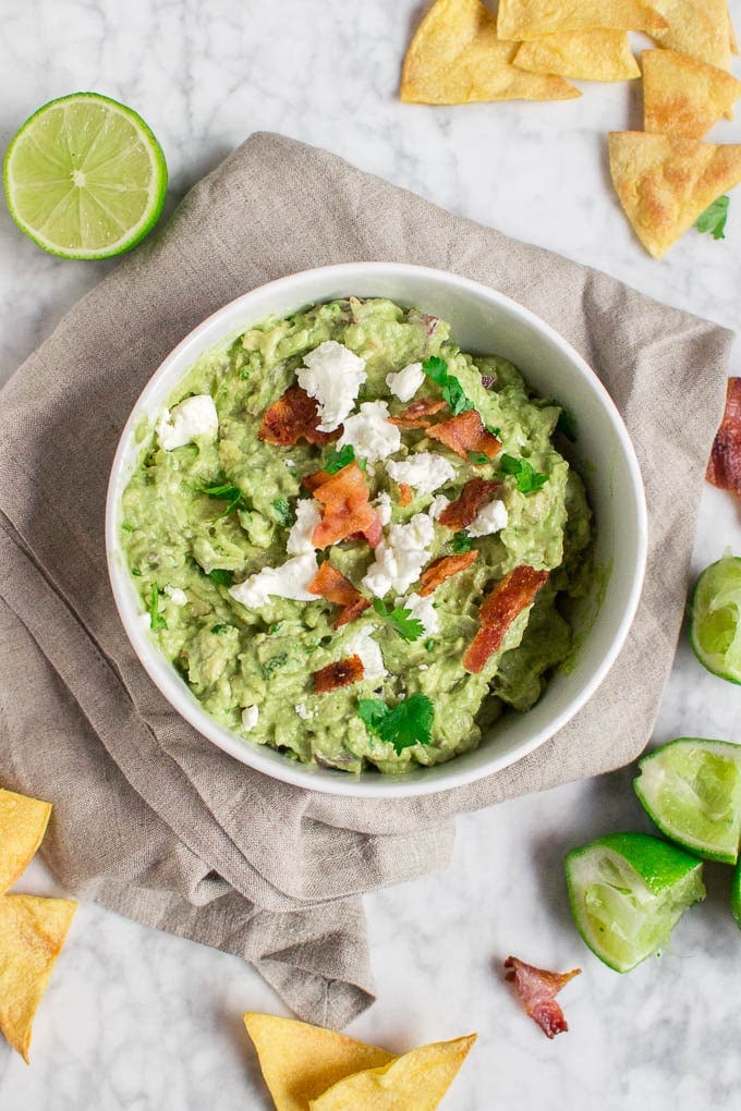 An aerial view of a bowl filled with goat cheese bacon guacamole topped with fresh cilantro on a marble table, surrounded by tortilla chips, fresh limes, and crumbled bacon.