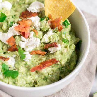 A bowl filled with guacamole, topped with bacon, goat cheese, cilantro and a tortilla chip.