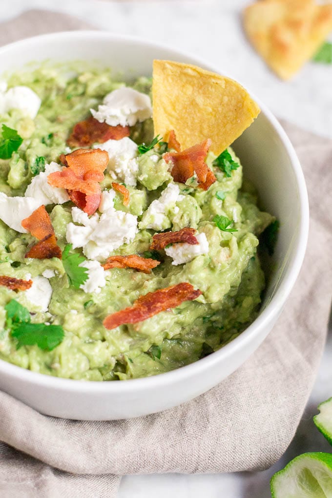 A bowl filled with guacamole, topped with bacon, goat cheese, cilantro and a tortilla chip. 