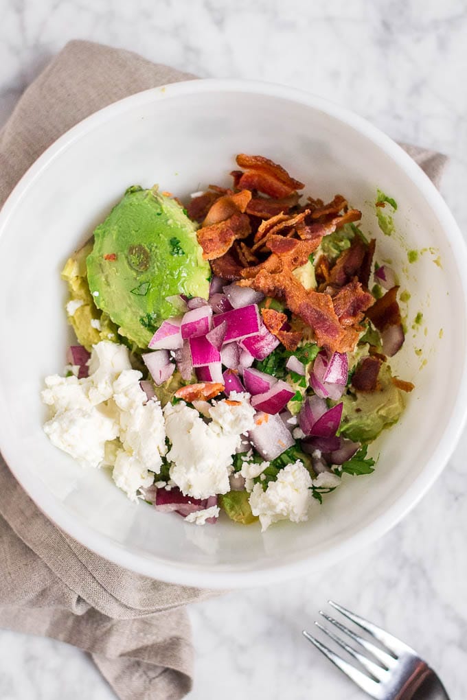 A white bowl filled with avocado, goat cheese, bacon, cilantro, jalapeno, lime juice, red onion and salt, with a fork. 