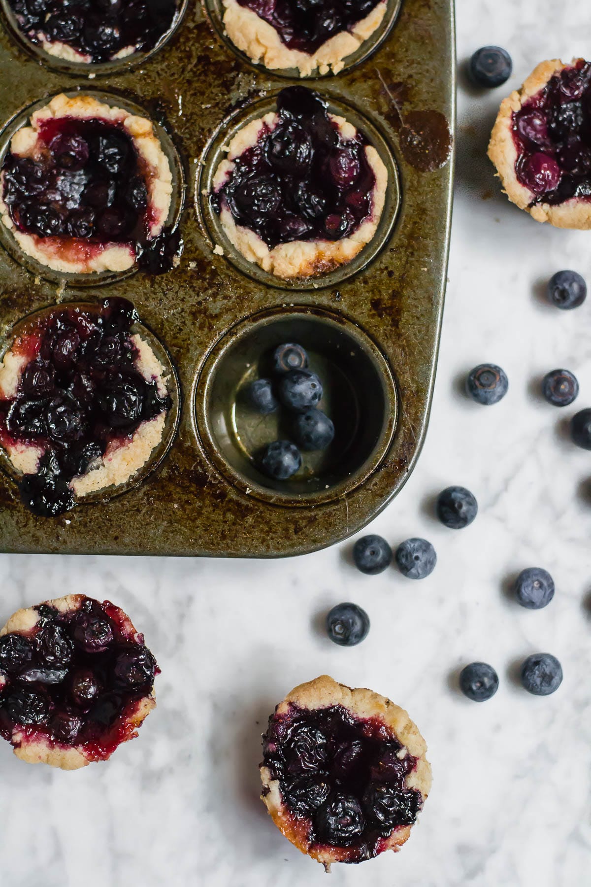 A mini muffin tin filled with mini blueberry tarts with fresh blueberries on a marble table. 