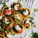 Aerial view of mini beef taco cups, topped with sour cream, tomato, cheddar cheese, green onion and cilantro on a striped napkin.