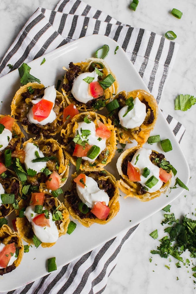 Aerial view of mini beef taco cups, topped with sour cream, tomato, cheddar cheese, green onion and cilantro on a striped napkin.