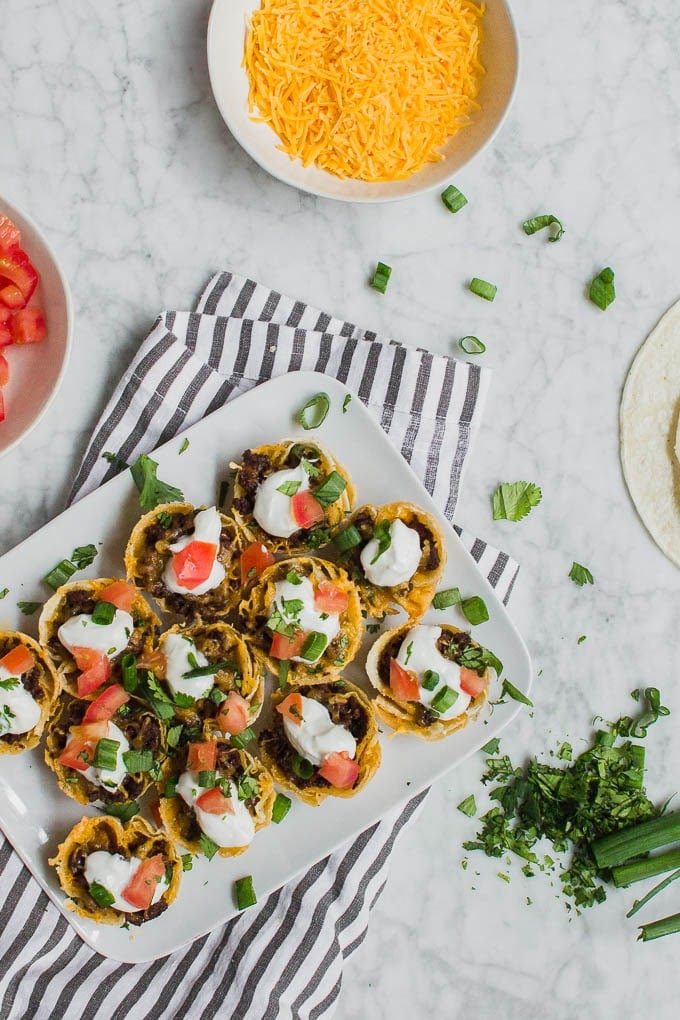An aerial view of mini beef taco cups on a white plate, surrounded by bowls of toppings - tomatoes, cheddar cheese, green onions, cilantro and some corn tortillas on a white marble table. 