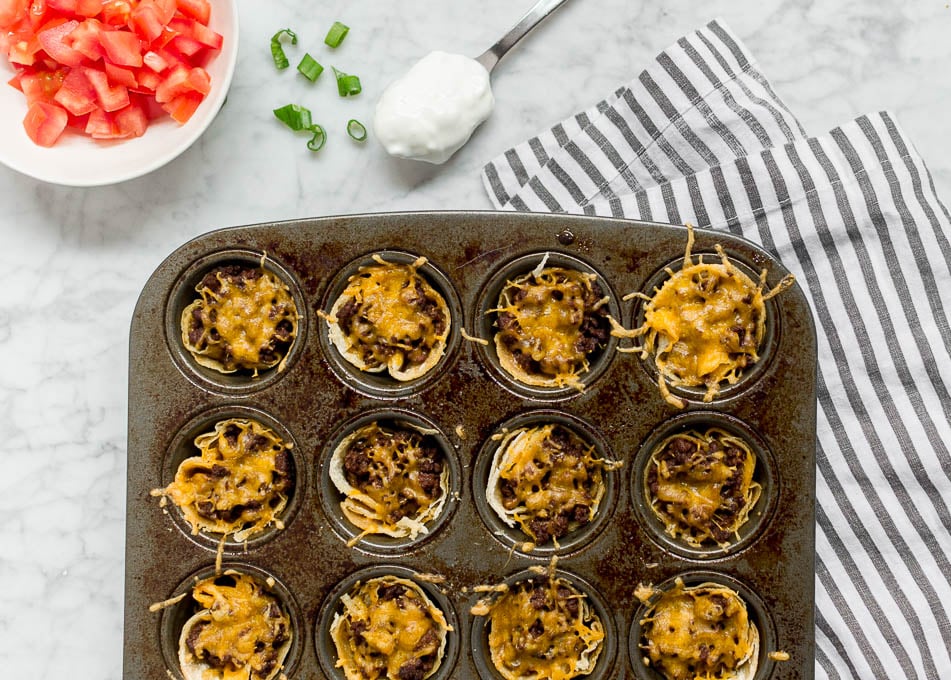 A mini muffin tin filled with corn tortillas, ground beef and cheddar cheese fresh from the oven. Surrounded by tomatoes, green onion and sour cream toppings. 