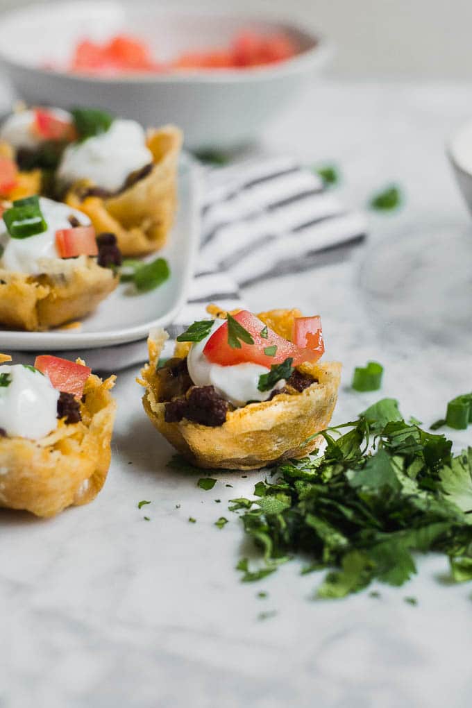 A few gluten-free mini taco bites with ground beef, sour cream, tomato, cheddar cheese on a marble table surrounded by toppings.