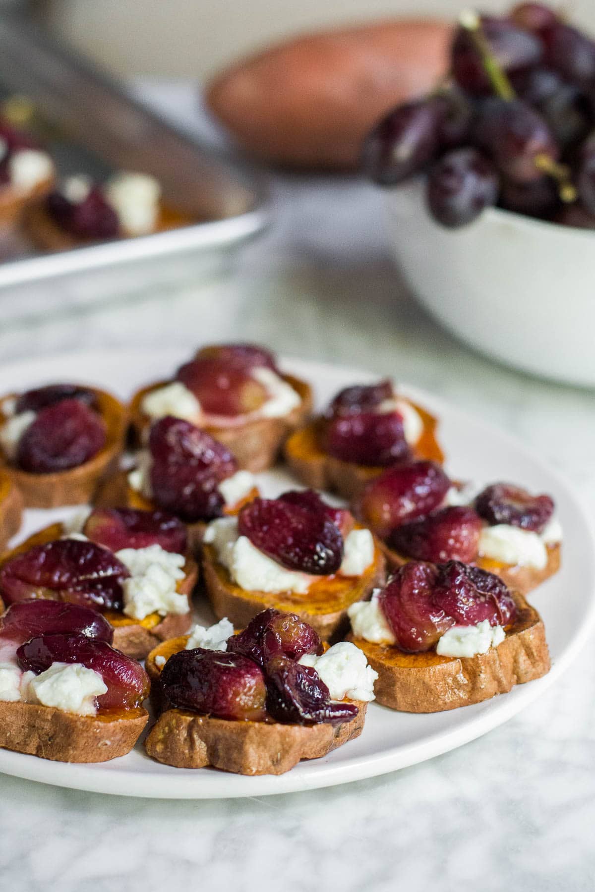 A plate of roasted sweet potato rounds topped with goat cheese, roasted grapes and honey, with a bowl of fresh grapes, a raw sweet potato and a pan filled with more sweet potato rounds. 