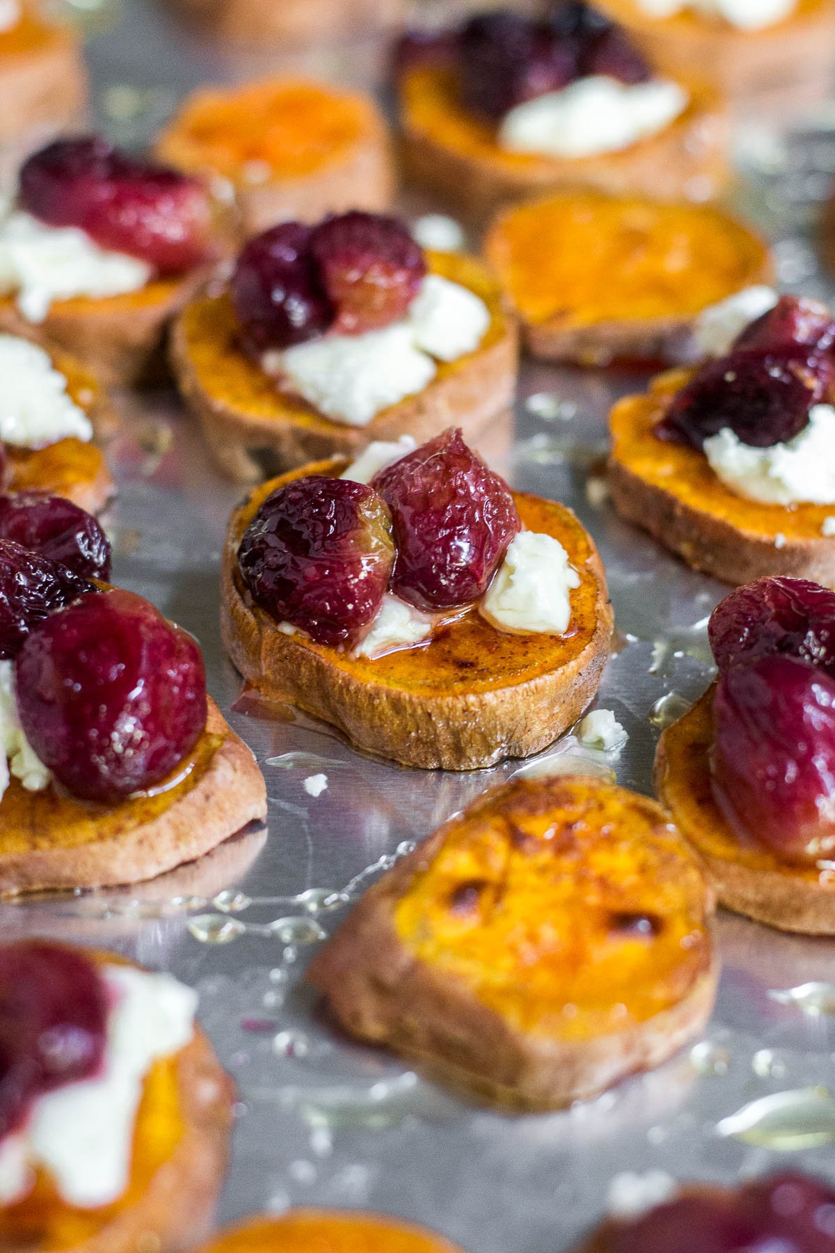 A baking sheet full of roasted sweet potato rounds topped with roasted grapes, crumbled goat cheese and honey.