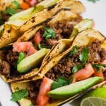 Aerial view of ground beef tacos in charred corn tortillas, sliced avocado, diced tomatoes, and chopped cilantro.