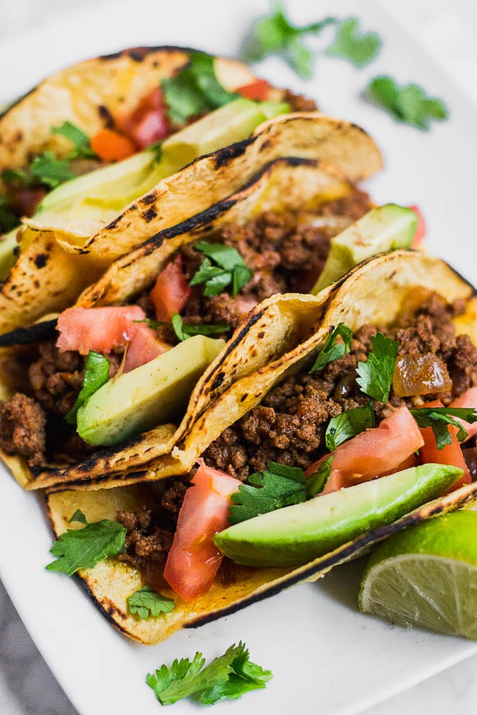Aerial view of ground beef tacos in charred corn tortillas, sliced avocado, diced tomatoes, and chopped cilantro.