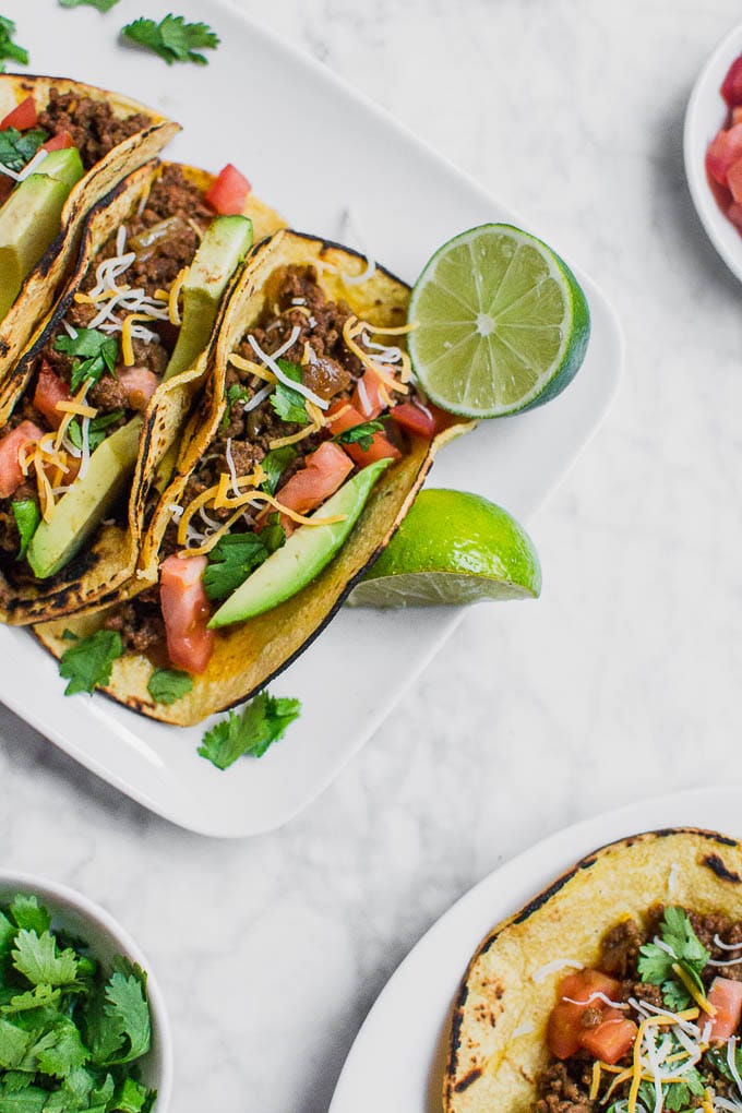 Aerial view of a plate of ground beef tacos with limes, a bowl of cilantro, tomatoes, and sliced avocado. 