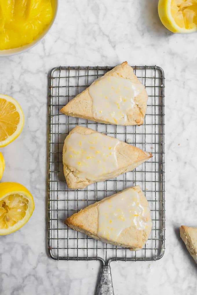 An aerial view of a rectangle baking cooling rack with three lemon scones with lemon glaze and a bowl of lemon curd, with lemon halves. 