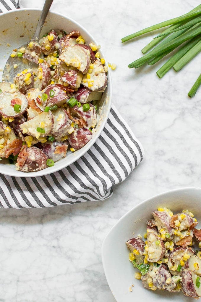 A large serving bowl filled with roasted corn, bacon potato salad with a serving spoon and a smaller bowl of a small portion of red potato salad. 