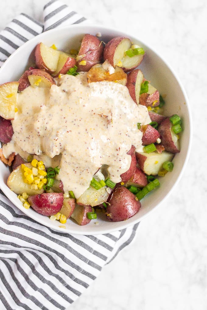 A large bowl with poatoes, corn, green onion and bacon with a creamy mustard sauce on top for potato salad. 