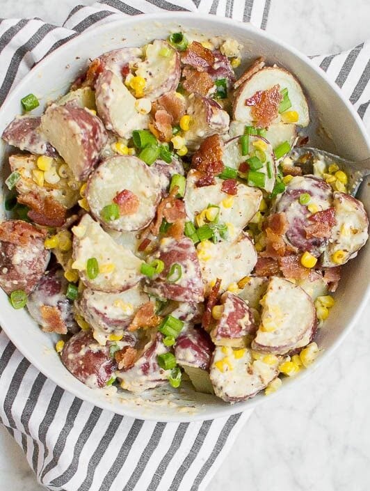 A bowl of Roasted Corn and Bacon Red Potato Salad with sliced red potatoes, corn, bacon bits, and chopped green onions sits on a striped cloth. A serving spoon rests in the bowl, with a plate and forks nearby.