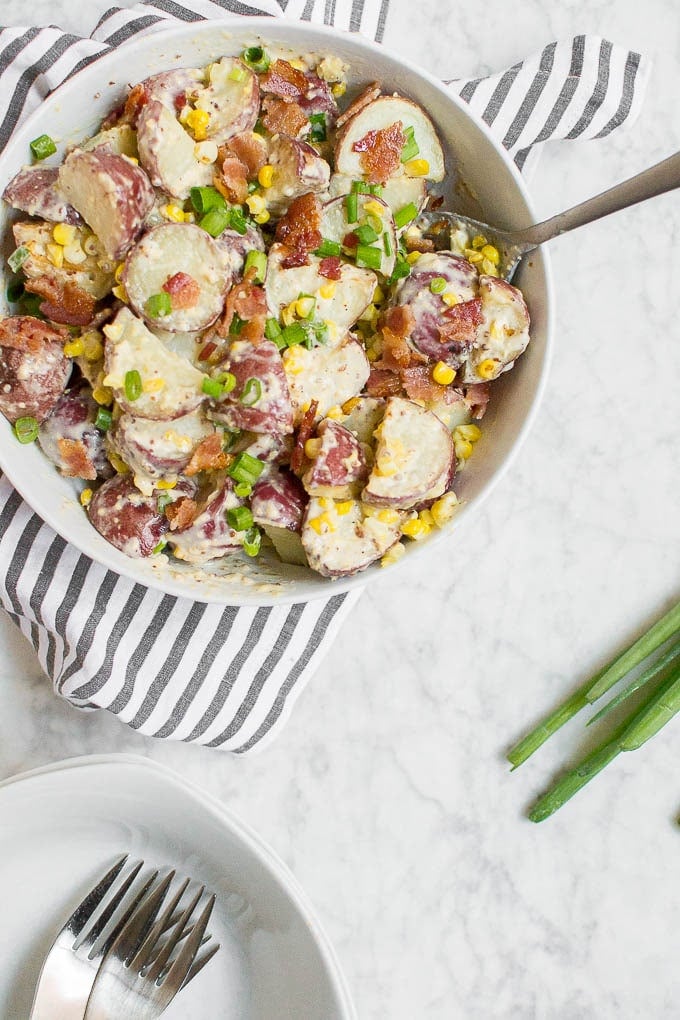 Aerial view of a large bowl filled with potato salad with corn, green onions, and bacon with a spoon. 