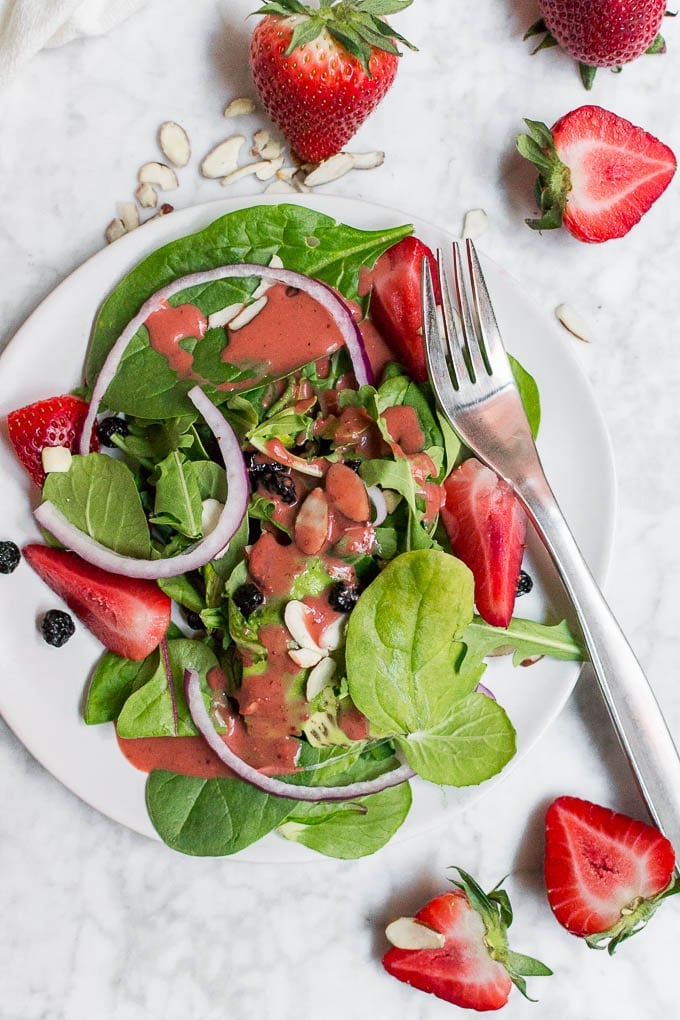 An aerial view of a plate of spinach salad with strawberries, red onion, dried blueberries and a fork. 