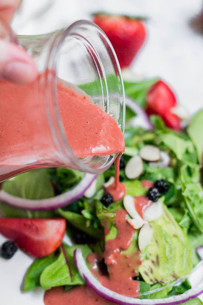 A cup of strawberry balsamic dressing being poured onto a strawberry spinach salad.