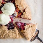 A rustic gluten-free vegan galette filled with blueberries, peaches, and rhubarb, topped with vanilla dairy-free ice cream with a wedge cut out of it on brown parchment paper.