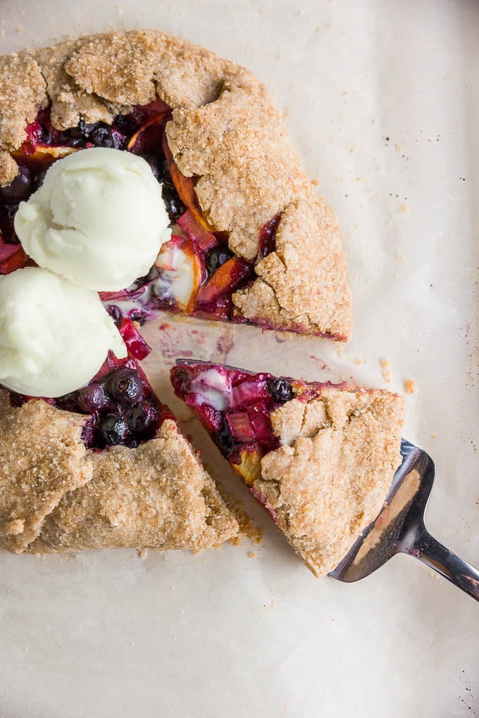 A rustic gluten-free vegan galette filled with blueberries, peaches, and rhubarb, topped with vanilla dairy-free ice cream with a wedge cut out of it on brown parchment paper. 