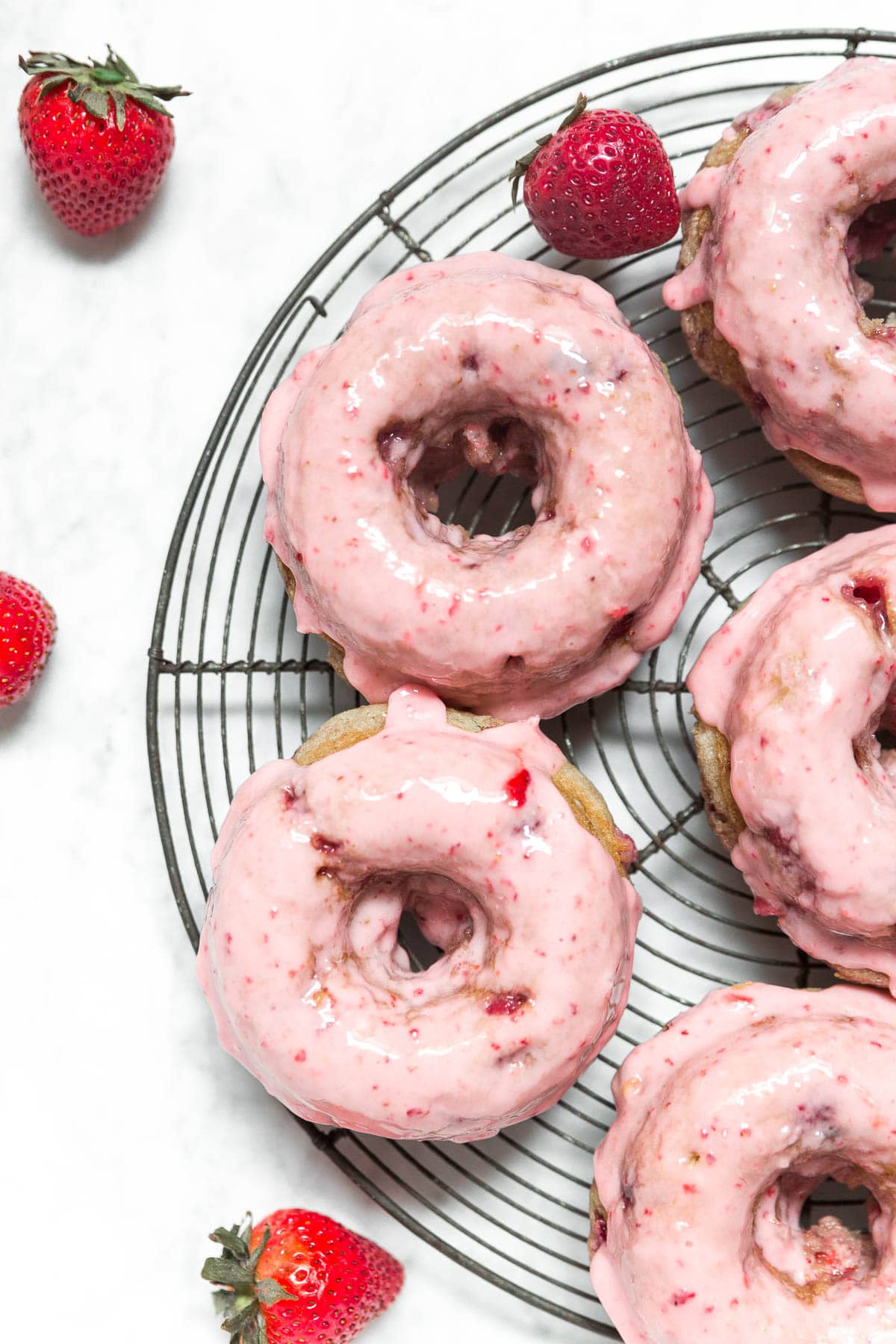Gluten-free baked strawberry donuts on a cooling rack and a cake stand with fresh strawberries and pink frosting.