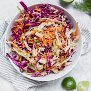 A white bowl containing cabbage dill slaw with red cabbage, green cabbage, carrots and dill with lime wedges on a table and fresh dill.