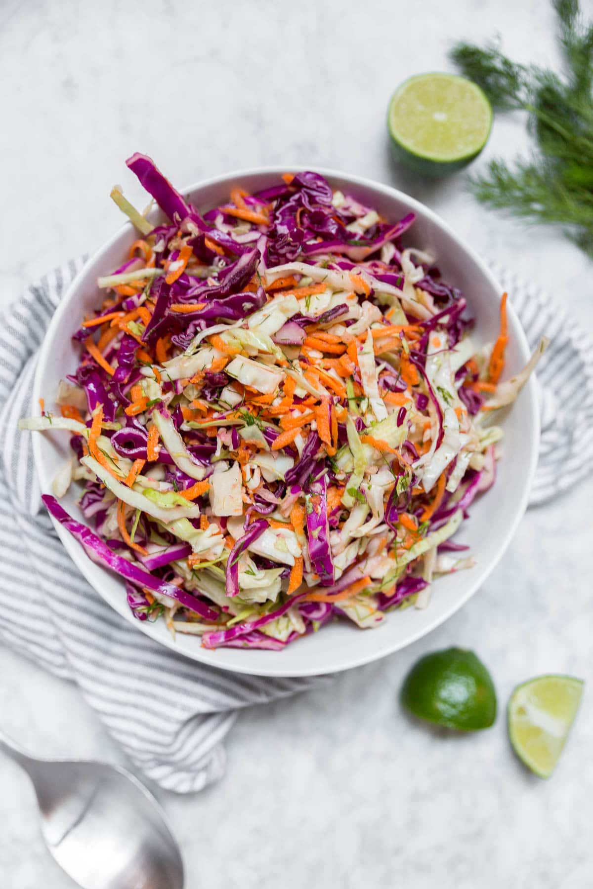 A white bowl containing cabbage dill slaw with red cabbage, green cabbage, carrots and dill with lime wedges on a table and fresh dill.