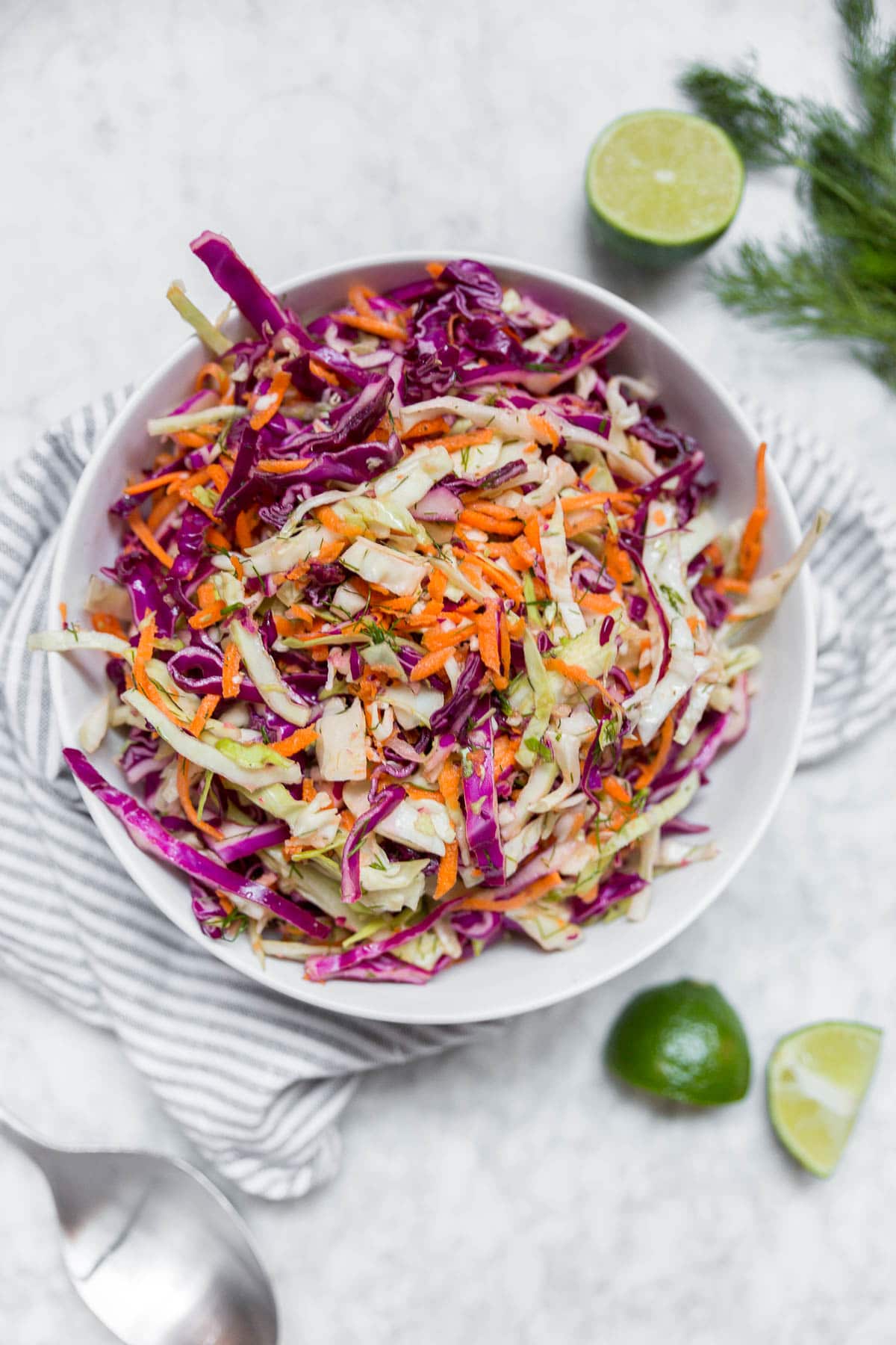 A white bowl containing cabbage dill slaw with red cabbage, green cabbage, carrots and dill with lime wedges on a table and fresh dill. 