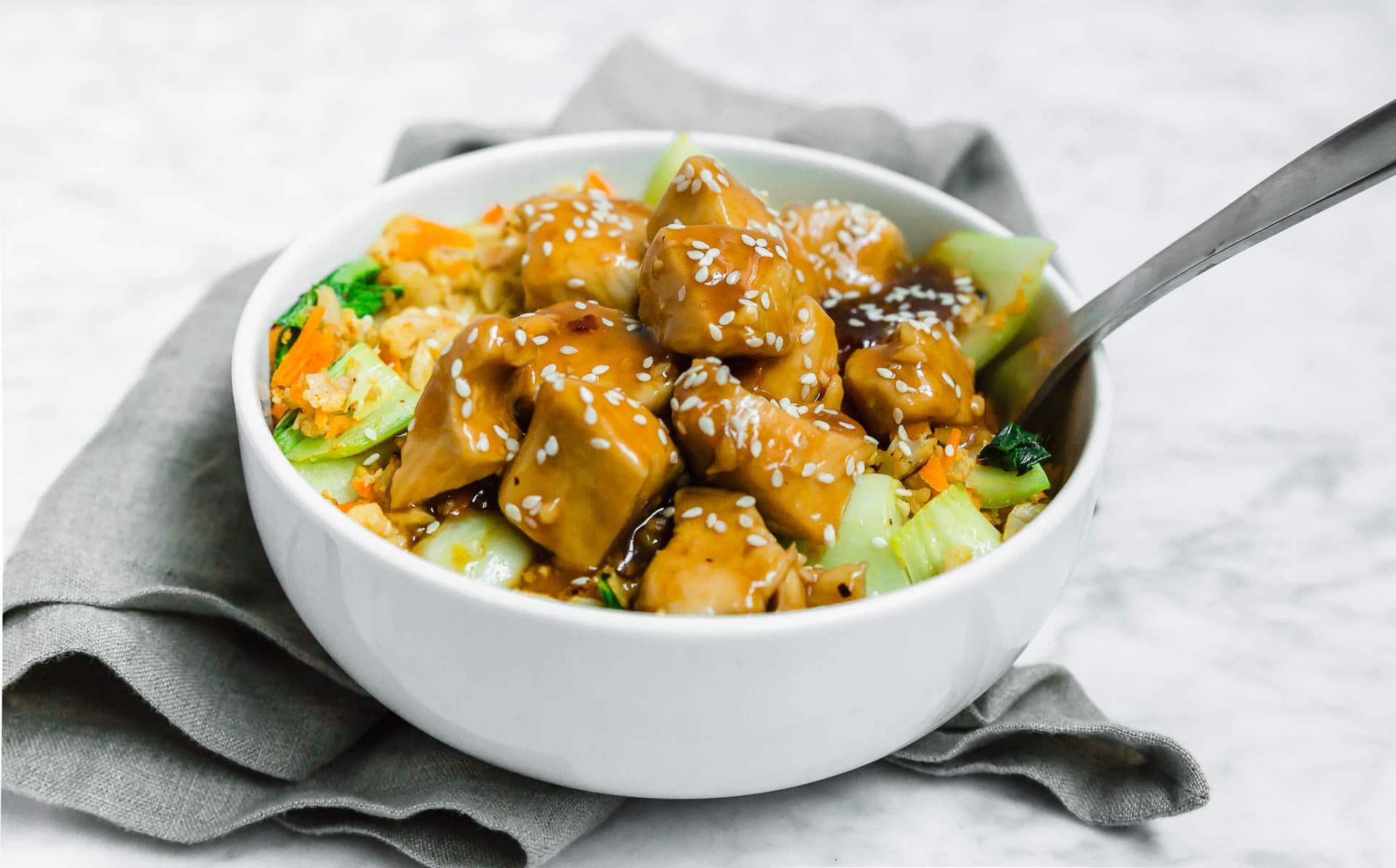 A straight on view of a white bowl with chicken teriyaki and cauliflower rice with bok choy with a spoon and gray napkin. 