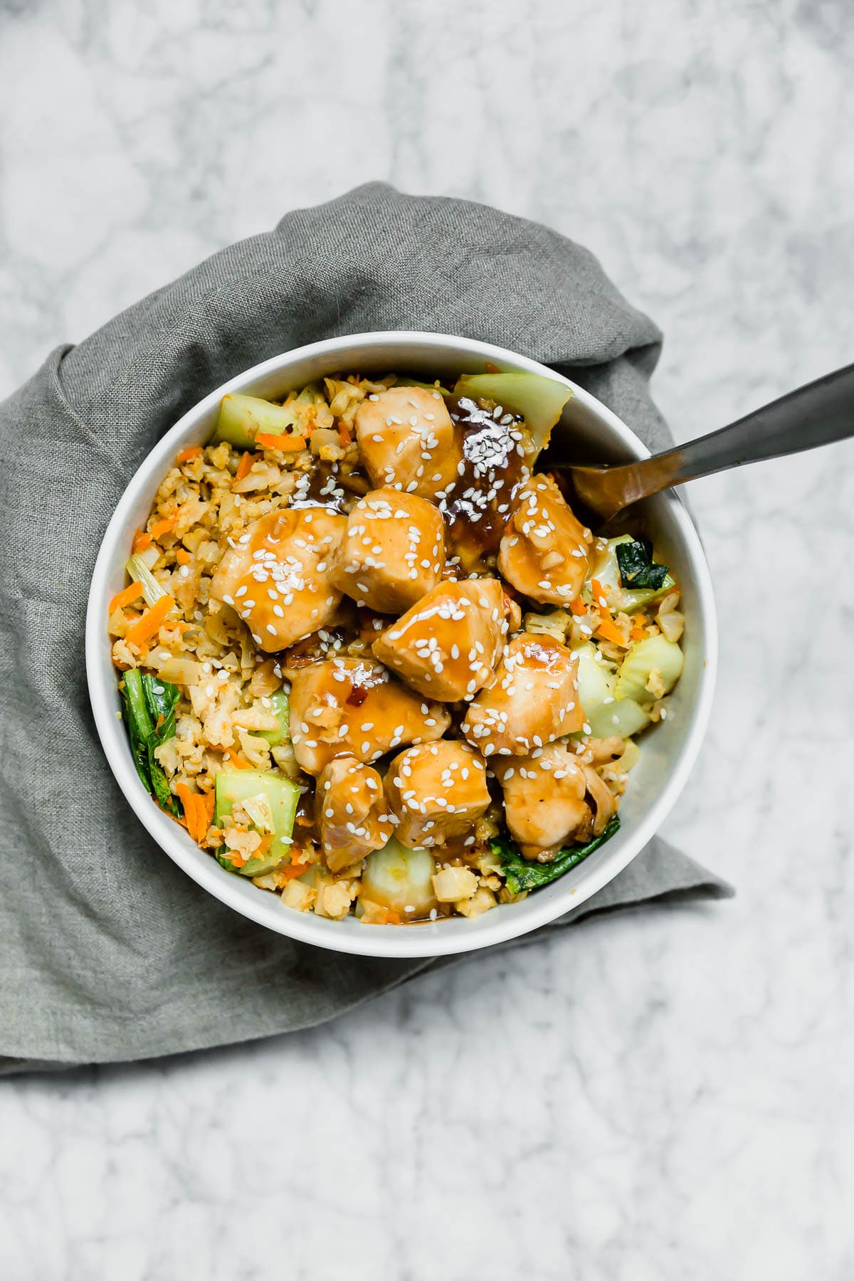 An aerial view of a bowl filled with paleo chicken teriyaki cauliflower rice with bok choy and carrots with a fork digging in and a gray napkin wrapped around the bowl. 