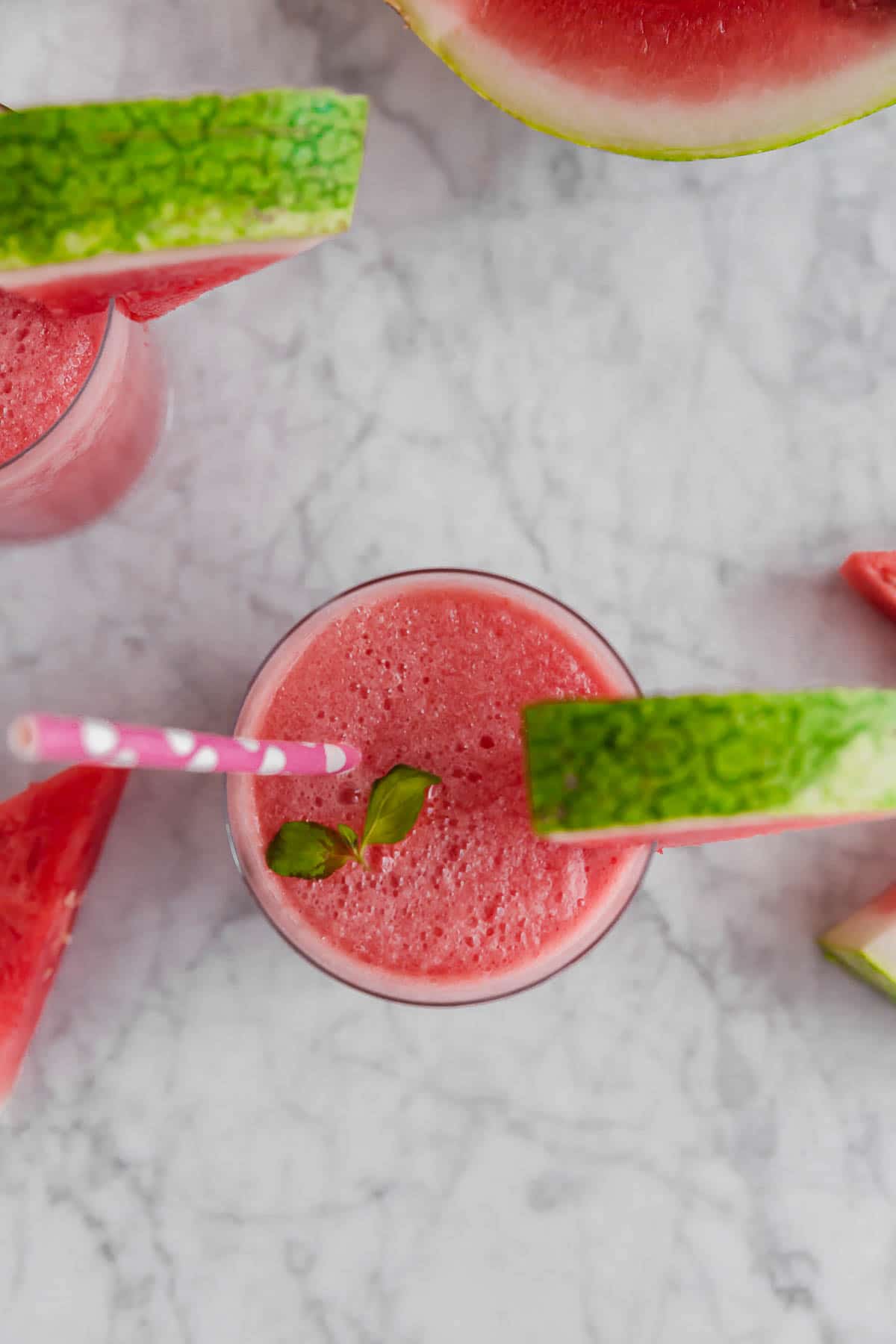 An aerial view of a glass with frozen watermelon rosé wine slushie with a watermelon wedge and pink straw and watermelon slices on a marble table.