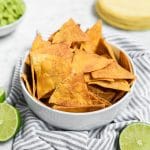 A white bowl filled with golden, crispy Baked Chili Lime Tortilla Chips sits on a striped cloth. Sliced limes and a stack of corn tortillas are nearby, with a bowl of guacamole in the background.