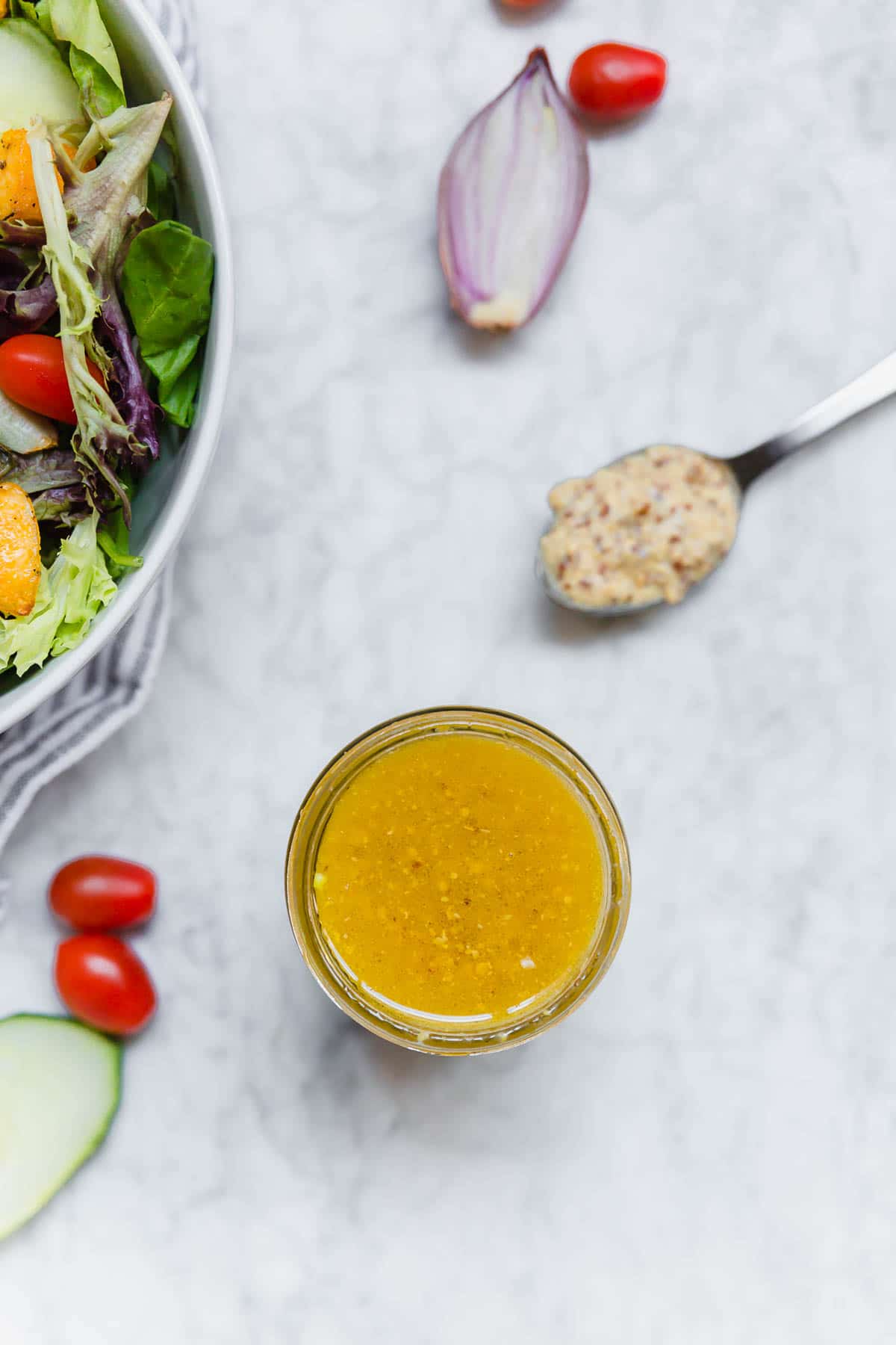 A Jar of honey mustard vinaigrette with a spoon of ground mustard, a salad, shallot and tomatoes. 