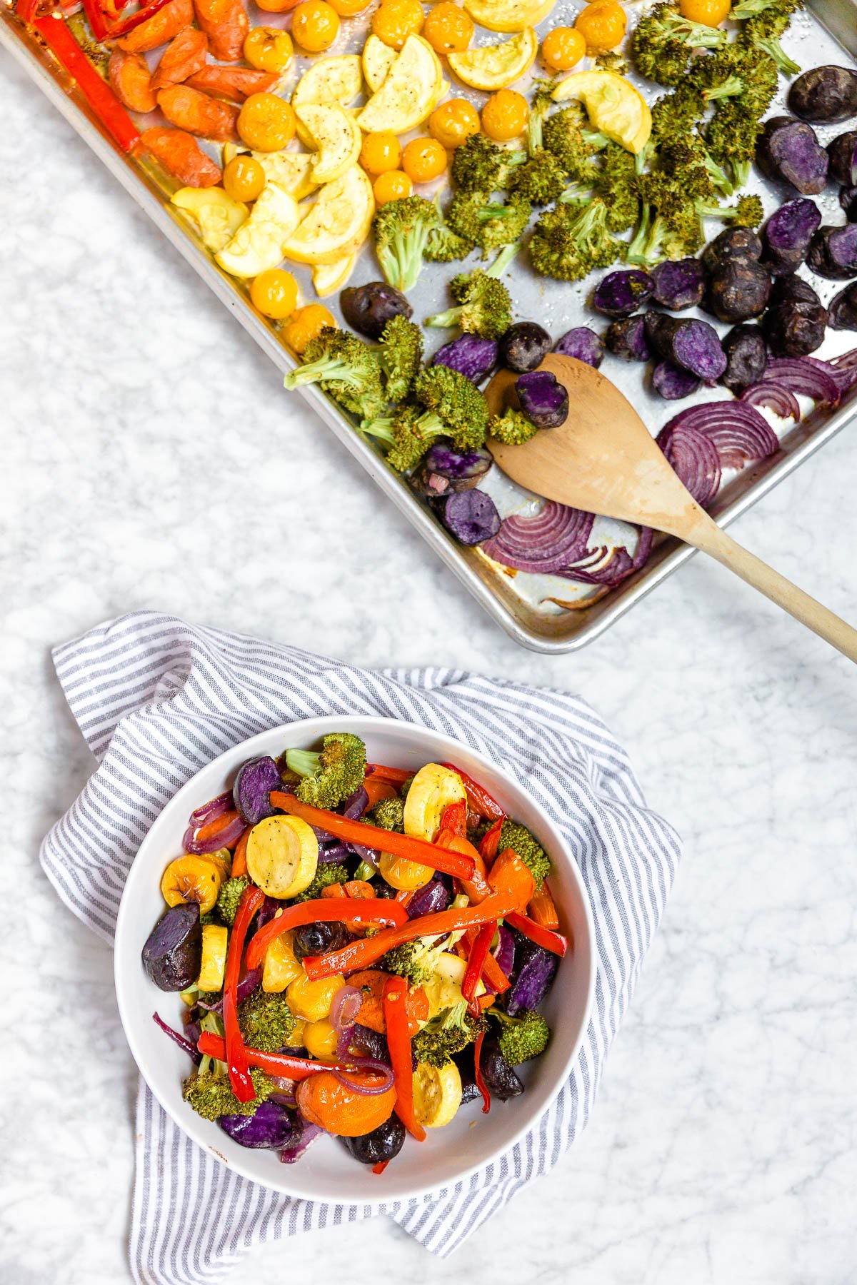 A sheet pan with rainbow vegetables that were roasted in the oven with a wooden spoon and a bowl with zucchini, tomatoes, broccoli, purple potatoes, bell peppers and carrots in it.