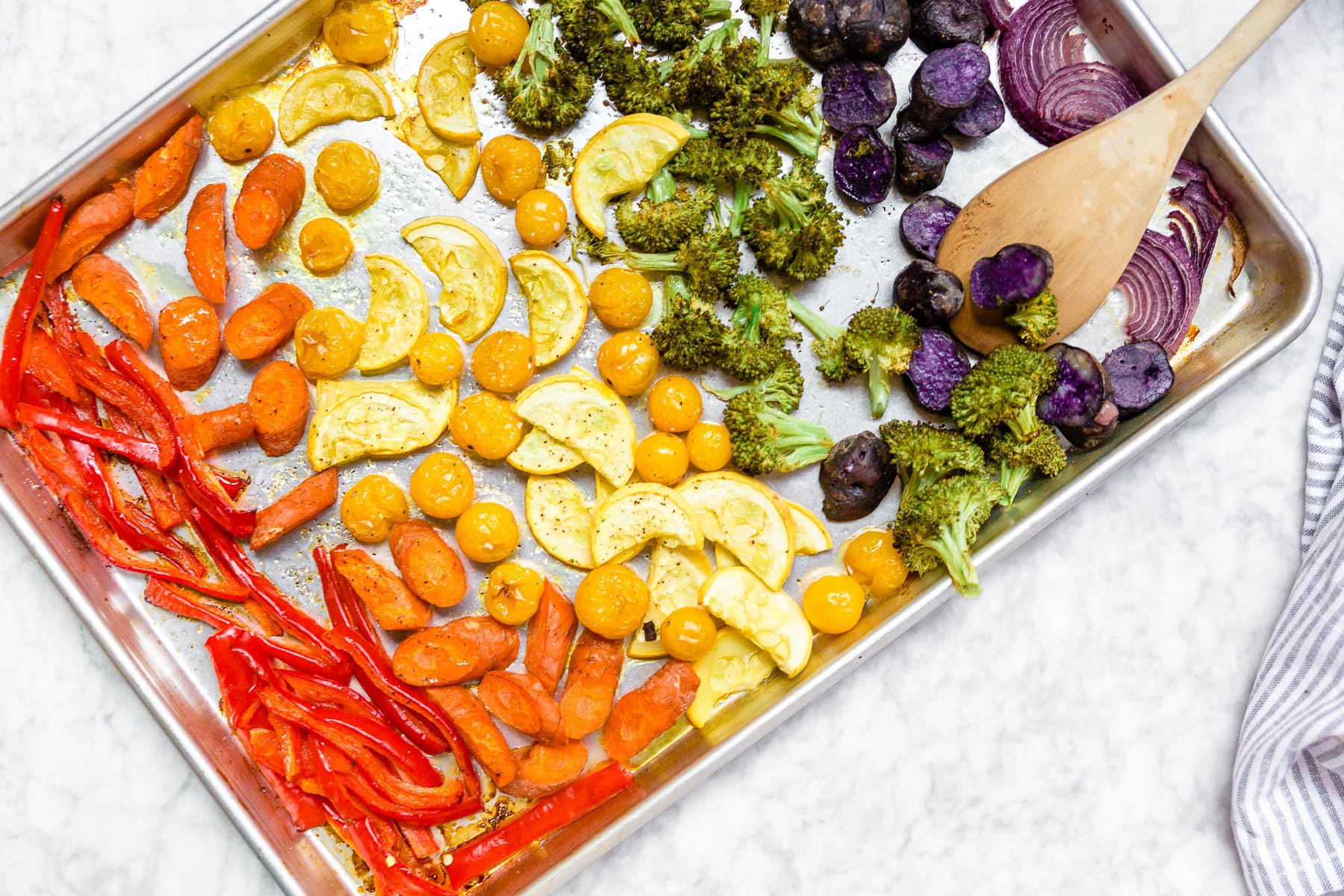 A sheet pan with roasted rainbow vegetables on it including red bell peppers, carrots, yellow squash, yellow tomatoes, broccoli, baby purple potatoes, and red onions with a wooden spoon.