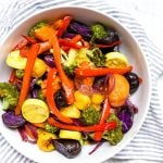 A bowl of mixed roasted vegetables in a rainbow of colors on a white table.