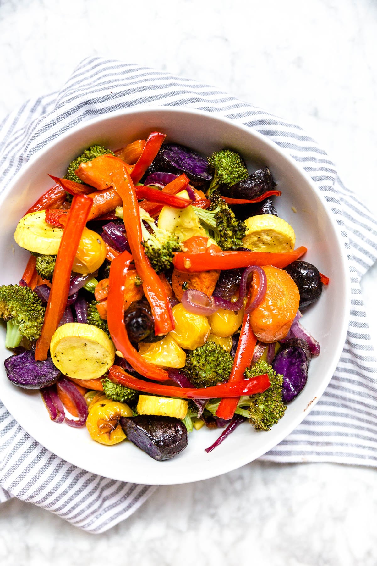 A bowl of mixed roasted vegetables in a rainbow of colors on a white table. 