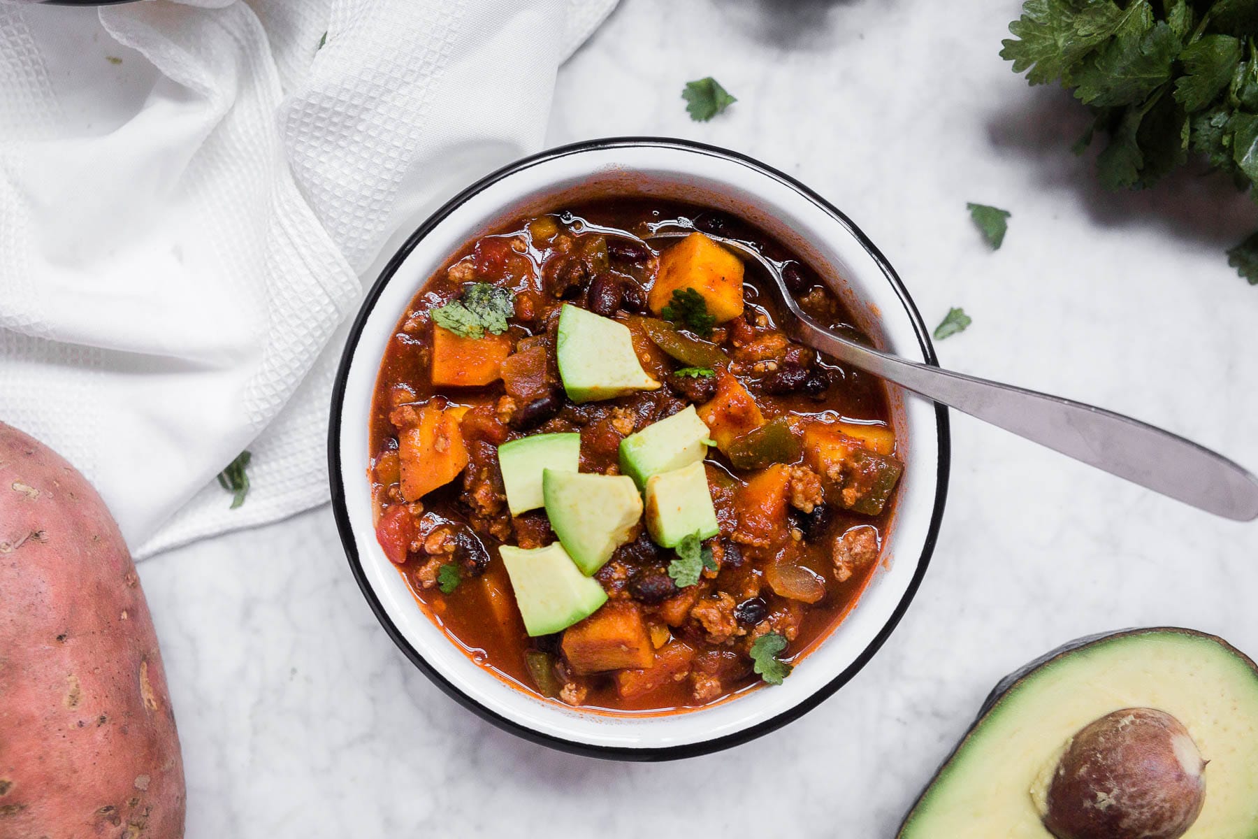 A bowl of slow cooker sweet potato turkey chili with avocado and cilantro and a silver spoon. 