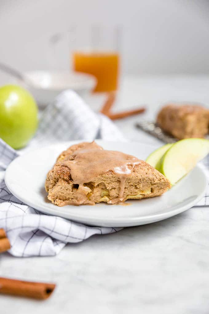 A plate with a gluten-free dairy-free apple cinnamon scone with cinnamon glaze with a glass of apple cider, sliced granny smith apples and cinnamon sticks. 