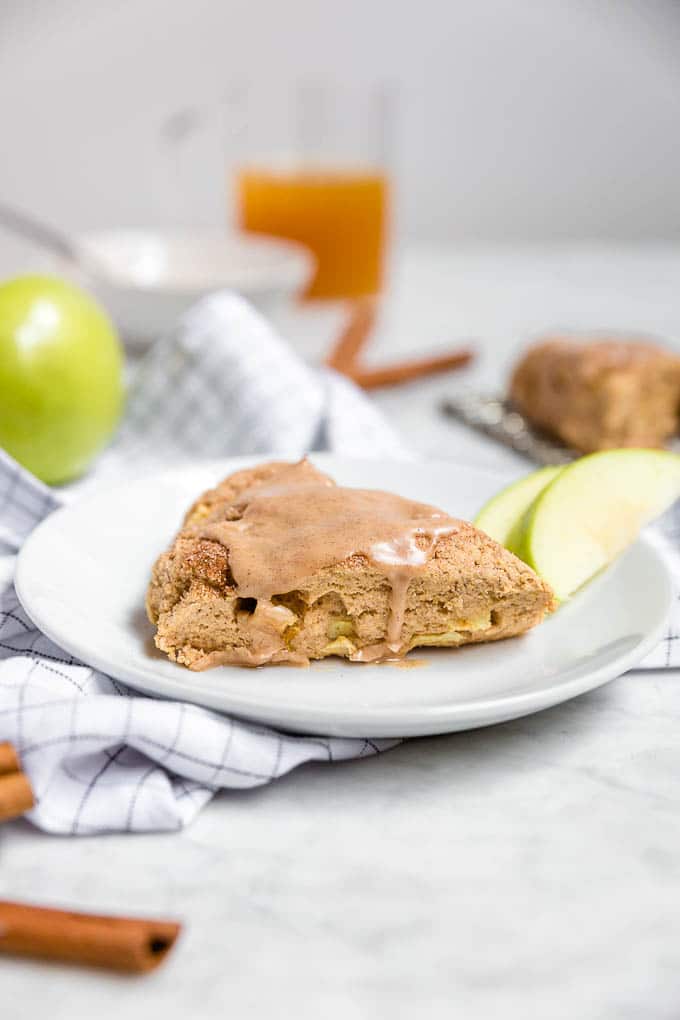 A plate with a gluten-free dairy-free apple cinnamon scone with cinnamon glaze with a glass of apple cider, sliced granny smith apples and cinnamon sticks.