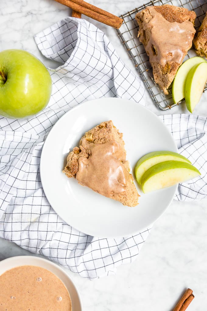 A plate with an apple cinnamon scone and slices of granny smith apple, a bowl of cinnamon apple glaze, and cinnamon sticks. 