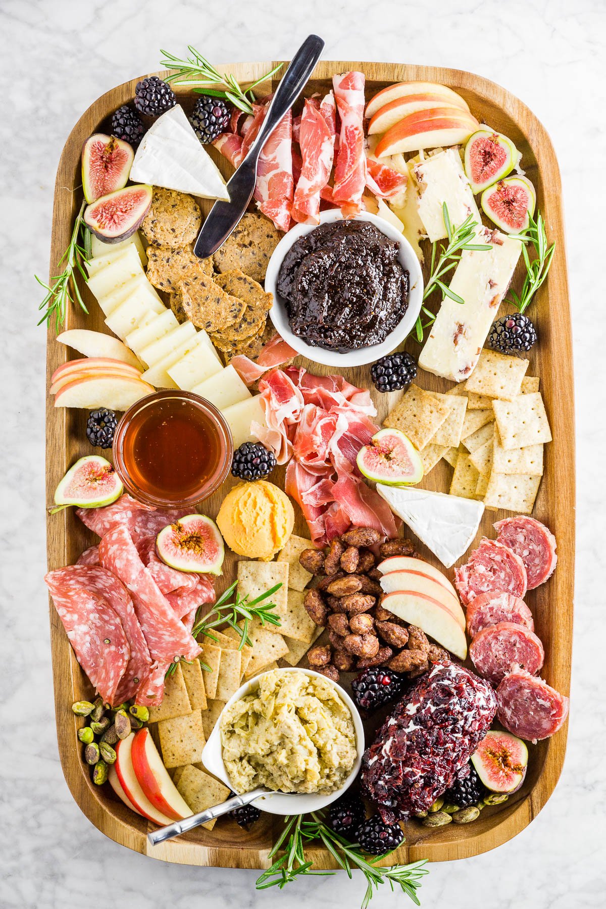 A wooden cheese board filled with cured meats, cheeses, apples, fig jam, berries and crackers. 