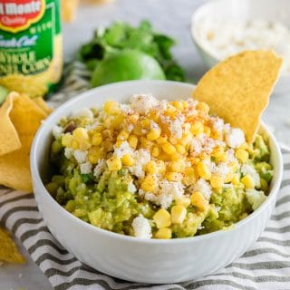 A bowl of guacamole with corn and cotija cheese with a corn tortilla chip in it.