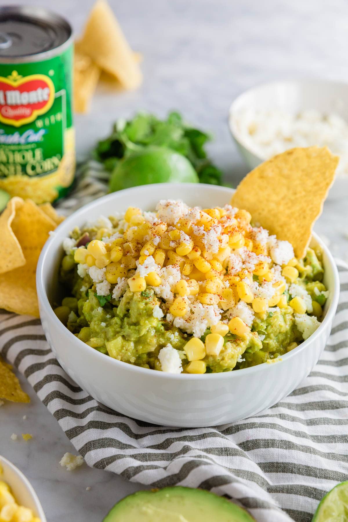 A bowl of guacamole with corn and cotija cheese with a corn tortilla chip in it. 