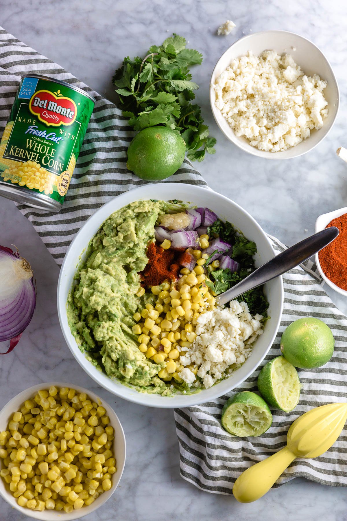 A bowl of mashed avocado with red onion, corn, cotija cheese, cilantro and paprika ready to be turned into corn and cotija cheese guacamole. 